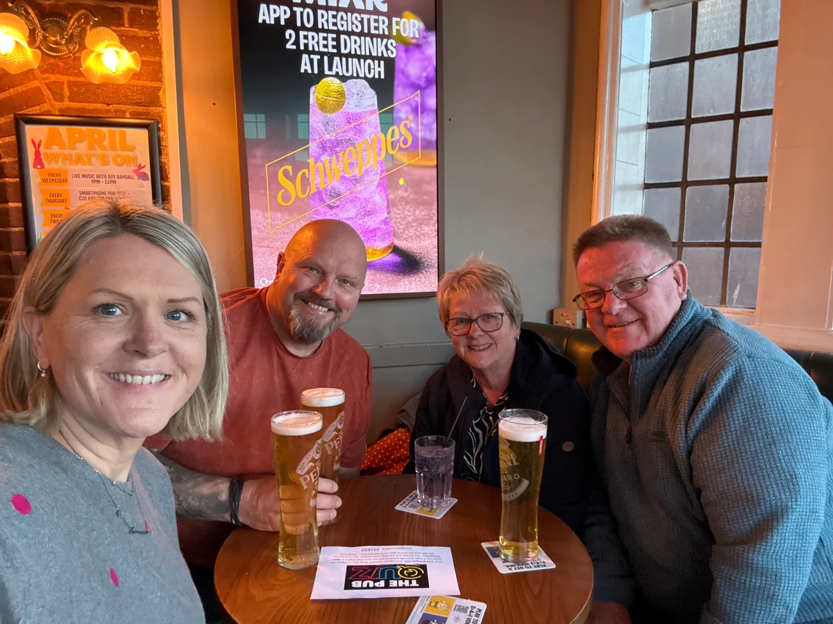 Four smiling teammates gathered around a pub table with a PubQuiz sheet